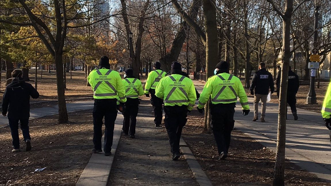 About 9 City Bylaw Enforcement, wearing high visibility vests, and corporate security walk through the park. One guard has papers in his hands. 